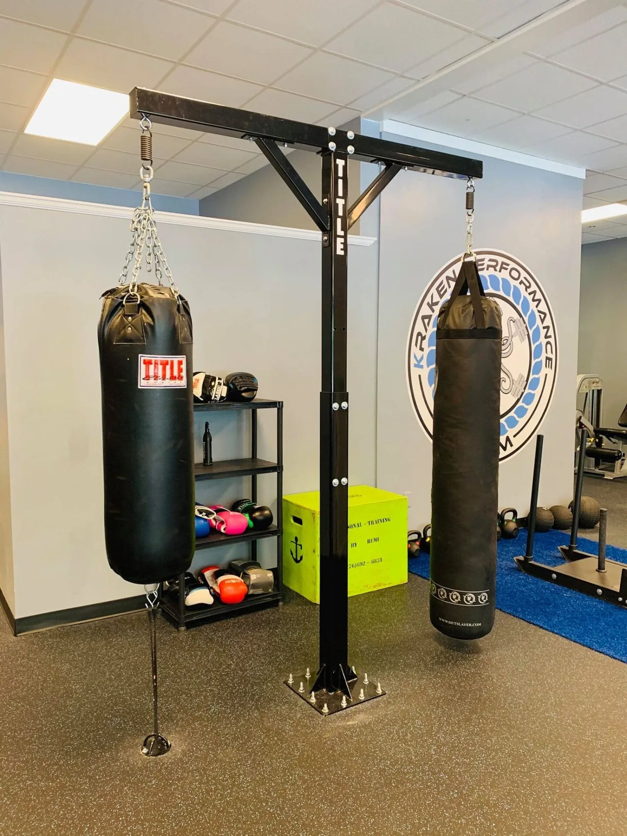 Heavy bags hanging from a TITLE rig in front of the painted Kraken Performance Gym wall logo, with kettlebells and the Personal-Training-By-Remi box.