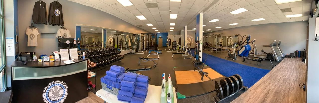 Panoramic view of Kraken Performance Gym floor: dumbbells, racks, and machines around a central blue runway.