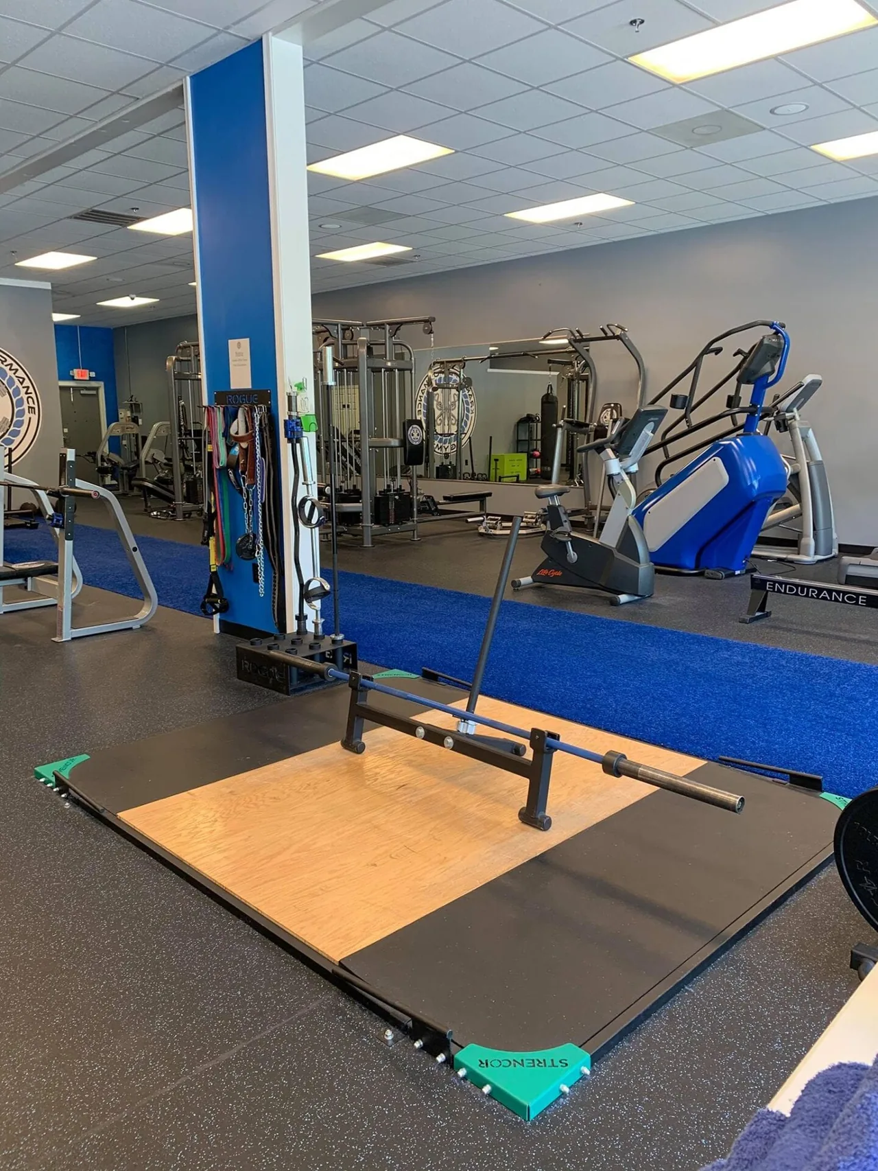 Strencor lifting platform with a loaded Olympic bar and bumper plates on a wood inset, mirrors and a recumbent bike behind.
