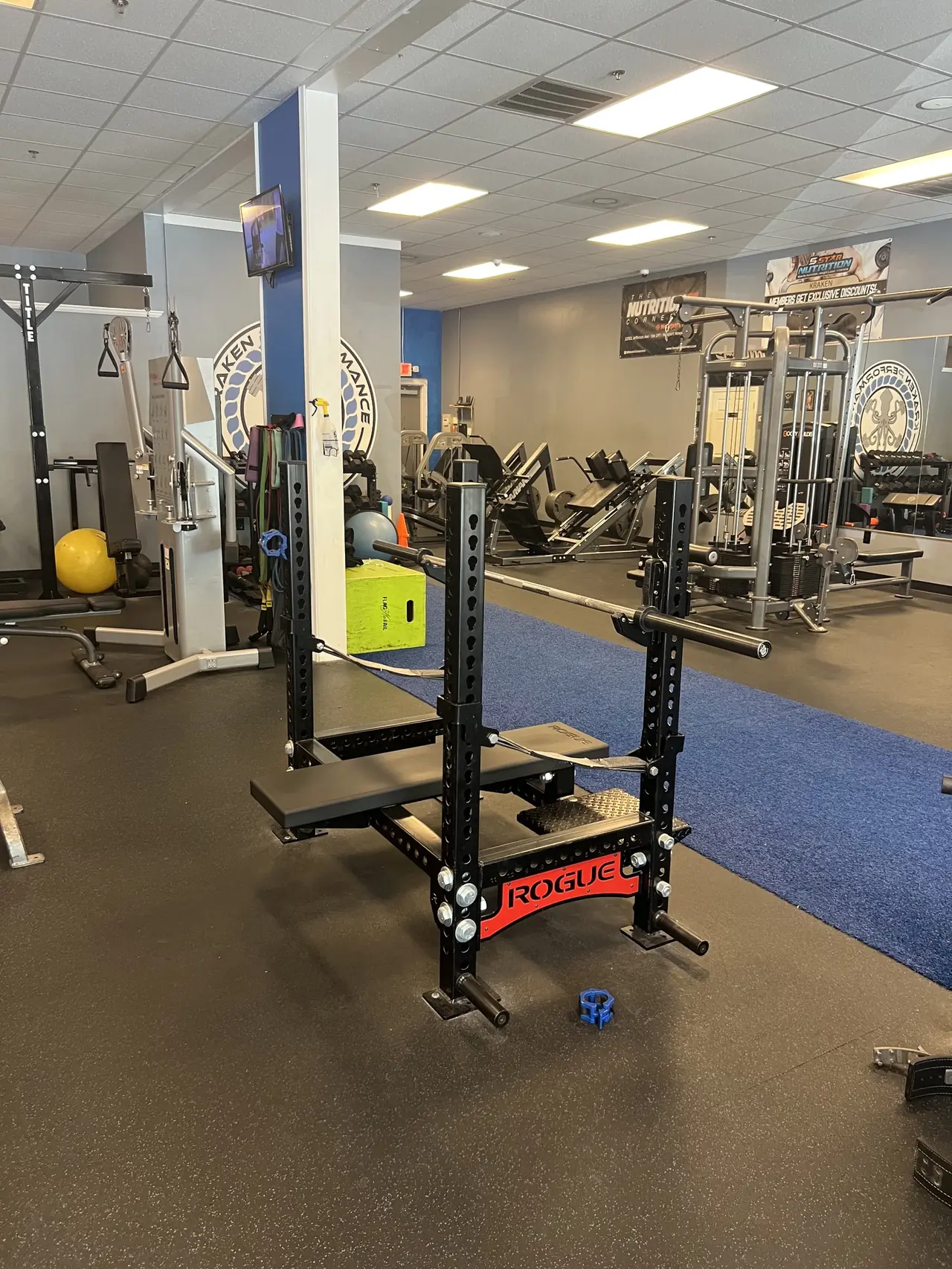 Rogue squat stand with a spotter deck on the open floor, cable stations and the blue turf runway behind.