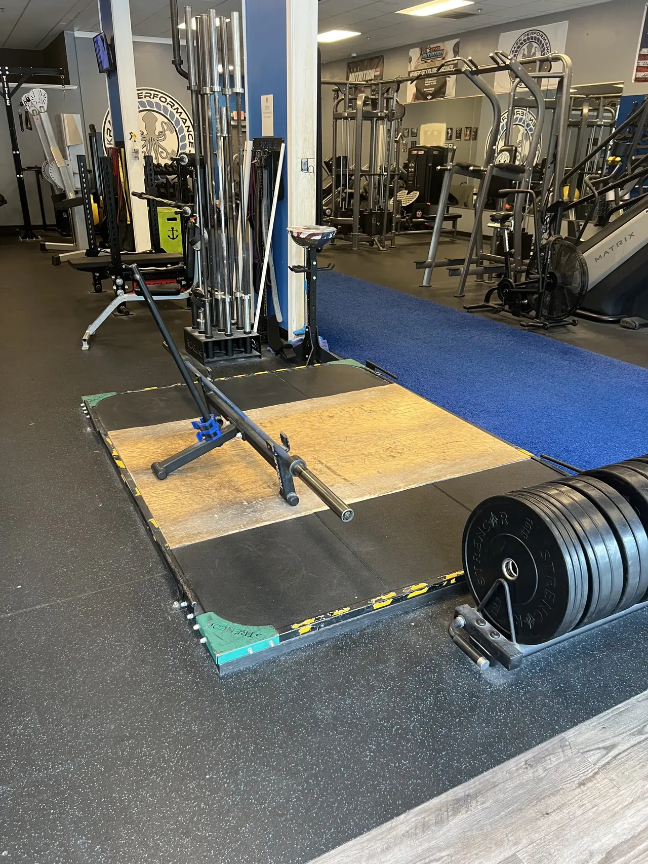 Strencor lifting platform with a bar on the jack, vertical barbell storage and a loaded plate cart alongside.
